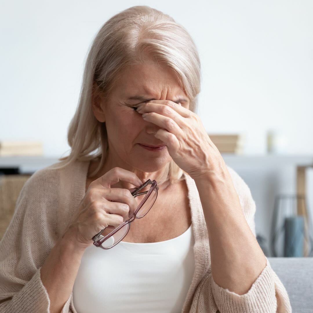 Older woman holding glasses in one hand and rubbing her eyes with the other hand